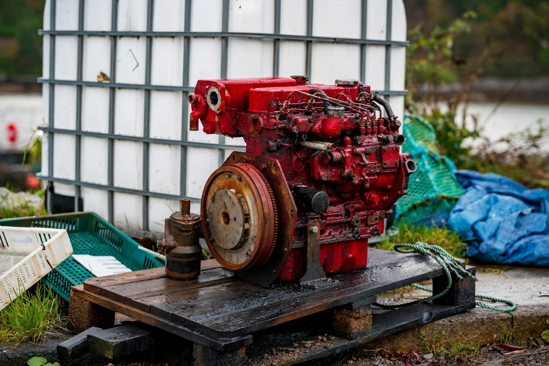 A rustic red diesel engine on a wooden pallet outdoors, surrounded by industrial objects.